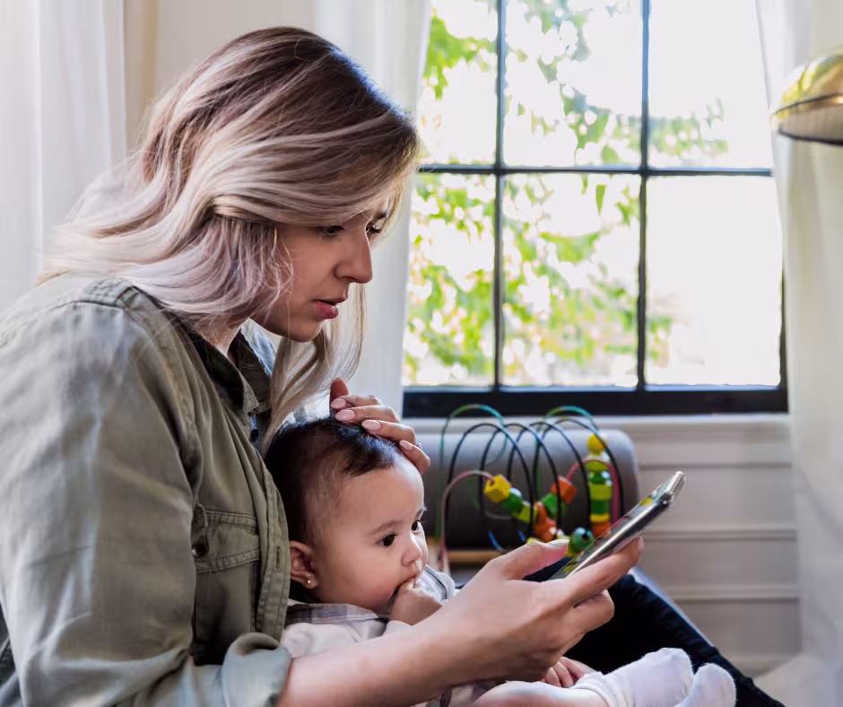 A woman seated, looking at a phone in her right hand with a baby in her lap and toy in the background